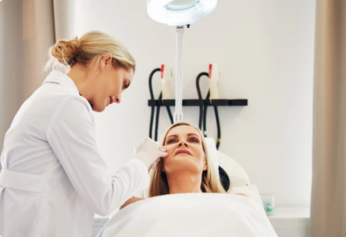 A female dermatologist wearing gloves examines a womans face in a well-lit clinic. The patient is lying back on a treatment chair under a bright examination lamp. Medical equipment is visible in the background.