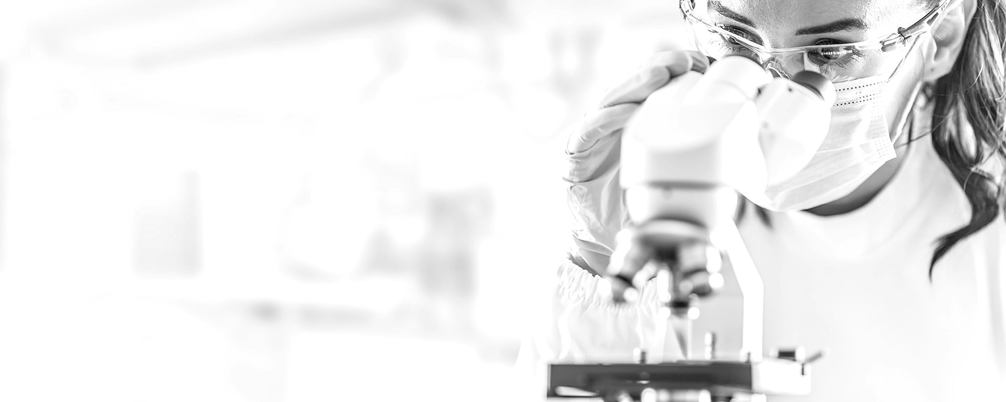 A scientist wearing safety glasses, a mask, and gloves looks into a microscope in a bright laboratory setting.