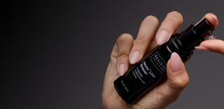A hand with manicured nails holds a black bottle of Revision Skincare Revox Line Relaxer, dispensing a small drop of the product from the pump against a dark gradient background.