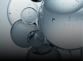 Close-up of transparent bubbles of various sizes clustered together, suspended in clear liquid against a gradient background that shifts from light at the top to dark at the bottom. Small air bubbles are visible inside the spheres.