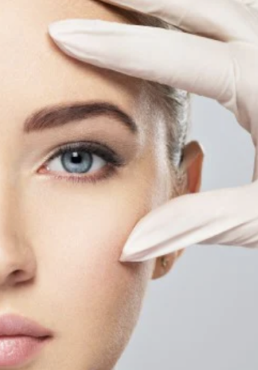 A close-up of a womans face, focusing on her blue eye. She is touching her temple and cheek with gloved fingers, suggesting a medical or cosmetic procedure. The background is neutral.