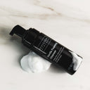 A black bottle of Revision Skincare Gentle Foaming Cleanser with a pump rests on a white surface, encircled by foam from this soap-free cleanser formulated with an amino acid surfactant.