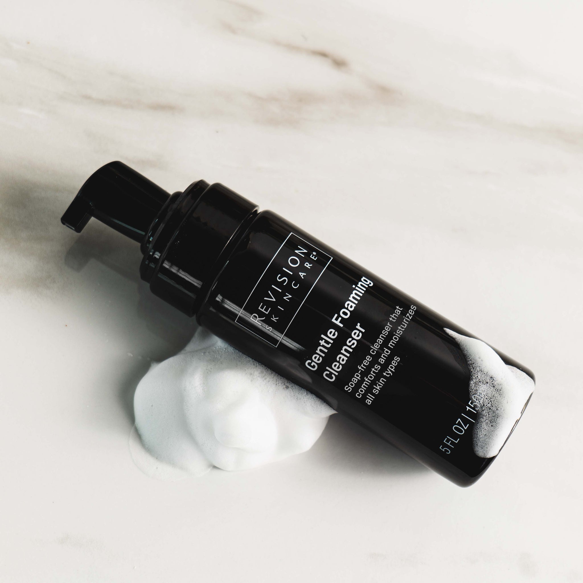 A black bottle of Revision Skincare Gentle Foaming Cleanser with a pump rests on a white surface, encircled by foam from this soap-free cleanser formulated with an amino acid surfactant.