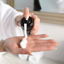 A person in a white robe dispenses Revision Skincare Gentle Foaming Cleanser from a black pump bottle onto their palm, with a mirror and towels in the background.