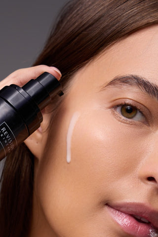 A close-up of a person applying a pump of white skincare serum to their cheek, holding a black bottle near their face. Their skin appears smooth and radiant.