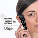A woman applies Revision Skincare C+ Brightening Eye Complex™ 0.5 oz under her right eye from a branded tube. Text reads: Reduce dark circles and under-eye puffiness with C+ Brightening Eye Complex™.