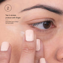 A close-up of a person gently tapping Revision Skincare’s C+ Brightening Eye Complex™ under their eye. On-screen text: “Tap in excess with finger. Avoid direct eye contact. Use twice daily to help reduce dark circles.”.