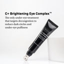 A tube of Revision Skincare C+ Brightening Eye Complex™ 0.5 oz, a brightening eye cream with a metal applicator tip, sits on a white surface by a cup. Text highlights its ability to target dark circles and under-eye puffiness.