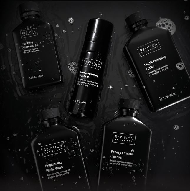 Five black bottles of Revision Skincare cleansing products are arranged on a wet black surface with water droplets and bubbles, including cleansing gel, foaming cleanser, cleansing lotion, facial wash, and enzyme cleanser.