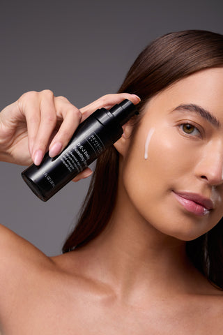 A woman applies a skincare serum to her face, holding a black bottle near her cheek, with a visible streak of product on her skin. She has smooth skin, straight brown hair, and a neutral background.