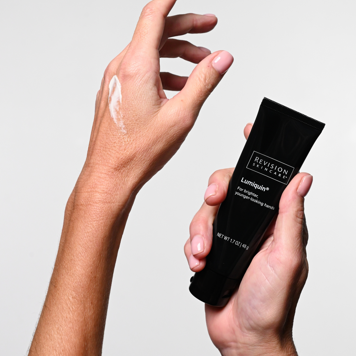 A person applies a white cream to their hand, holding a black tube labeled Revision Skincare Lumiquin® 1.7 oz—an age-defying moisturizer—against a plain white background.