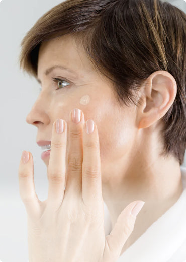 A woman with short brown hair applies a small amount of cream or foundation to her cheek, using her fingers. She is shown in profile against a light background.