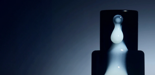 A close-up of a dark cylindrical bottle with a glossy surface, featuring a dramatic reflection of white liquid resembling a droplet, set against a gradient blue-black background.