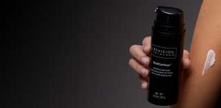 A hand holds a black bottle of Revision Skincare BodiContour next to a persons arm, which has a dab of white lotion on the skin, against a dark background.