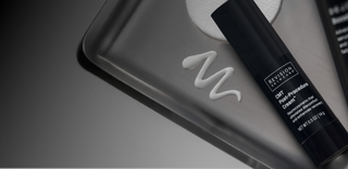 A black tube labeled Revision Skincare CMT Post-Procedure Cream lies on a metal tray with a wavy line of white cream next to it. The image has a gray gradient background.