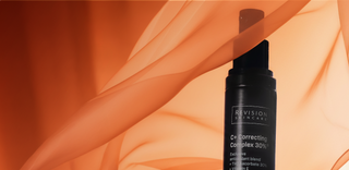 A black bottle of Revision Skincare C+ Correcting Complex 30% stands against a soft, flowing orange fabric background, partially obscured by the fabric’s translucent folds.