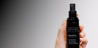 A hand with nude-colored nails holds a black bottle of Revision Skincare Nectifirm Advanced against a gradient gray background.