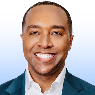 A man with short hair, wearing a teal suit jacket and white shirt, smiles broadly at the camera against a light blue and white gradient background.