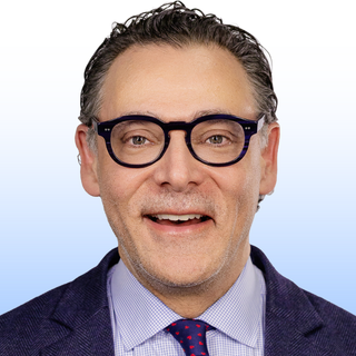 A man with short dark hair, wearing black glasses, a blue suit jacket, a light blue dress shirt, and a red and blue polka dot tie, smiles at the camera against a light blue gradient background.