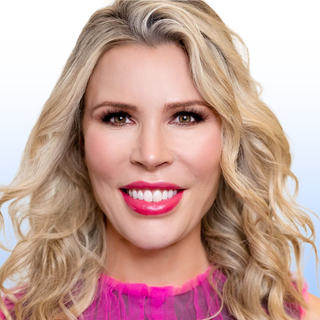 A woman with long, wavy blonde hair smiles at the camera. She is wearing bright pink lipstick and a sheer pink top, set against a light gradient background.