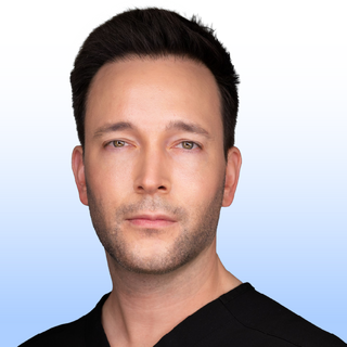 A man with short dark hair and light skin looks directly at the camera with a neutral expression. He is wearing a black shirt and the background is a light gradient from white to pale blue.