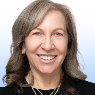 A smiling woman with shoulder-length gray hair, wearing a black top, layered necklaces, and hoop earrings, poses against a light gradient background.