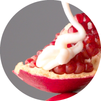 A close-up of a pomegranate half with white cream being drizzled over its red seeds, set against a gray background.