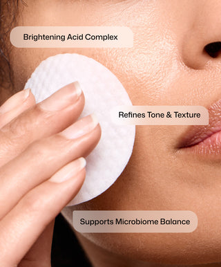 A person uses Revision Skincare’s Tone+ Correcting Pads™ on their cheek, gently applying brightening skincare to help address uneven skin tone. Text on the image reads: Brightening Acid Complex, Refines Tone & Texture, and Supports Microbiome Balance.