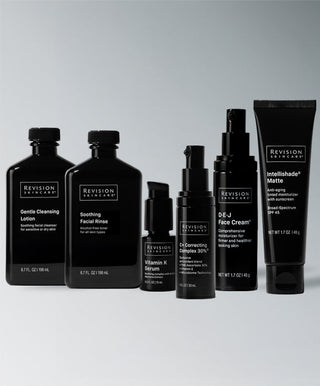 Seven black containers of Revision Skincare’s Anti-Redness Complete Full-Size Regimen are arranged in a row on a gray background, featuring cleanser, rinse, serums, creams, and sunscreen for sensitive and redness-prone skin.