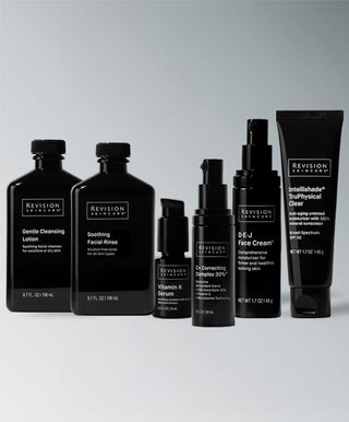 Six black bottles and tubes from Revision Skincare’s Anti-Redness Complete Full-Size Regimen are arranged in a row against a light grey background, each labeled for different steps, ideal for sensitive skin and anti-redness care.
