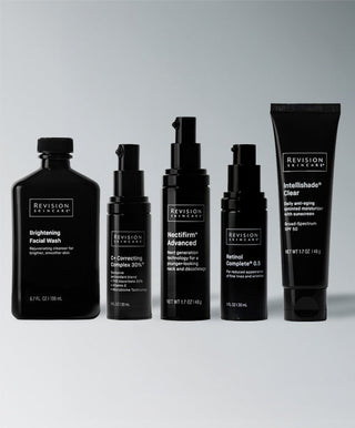 Five black bottles and tubes of Revision Skincare’s Brightening Full Size Regimen are neatly arranged in a row against a light gray background, showcasing the complete set for an effective brightening skincare routine.