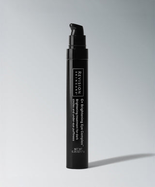 A sleek black pump bottle labeled "Revision Skincare C+ Brightening Eye Complex™," a brightening eye treatment, stands upright on a light gray background, casting a soft shadow to the right.