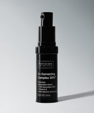 A black bottle of Revision Skincare's C+ Correcting Complex 30%® serum with a pump dispenser and white label highlighting key ingredients stands upright against a neutral background.