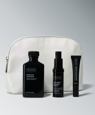 Against a gray gradient backdrop, the Revision Skincare Glow Essentials Gift Set features a white cosmetic bag with three black-labeled products: a brightening exfoliating cleanser, an energy-boosting serum, and a facial wash.