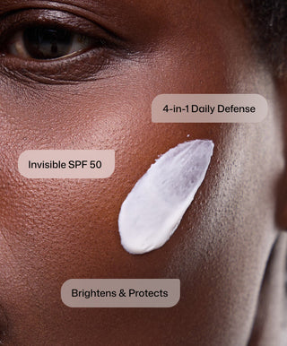 A close-up of a person with dark skin shows a swatch of Revision Skincare Intellishade® Clear, labeled Invisible Broad-Spectrum SPF 50. Text reads 4-in-1 Daily Defense and Brightens & Protects; it's used as an anti-aging moisturizer.