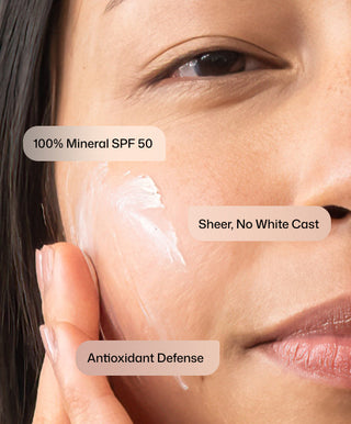 A woman applies Intellishade® TruPhysical Clear by Revision Skincare to her cheek, with labels reading 100% Mineral SPF 50, Sheer, No White Cast, and Antioxidant Defense.