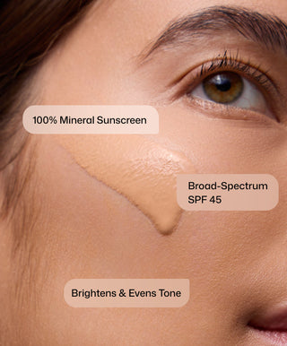 Close-up of a person’s cheek with a swipe of Intellishade® TruPhysical by Revision Skincare. Text reads: “100% Mineral Sunscreen,” “Broad-Spectrum SPF 45,” “Brightens & Evens Tone.” An anti-aging daily moisturizer for protection.