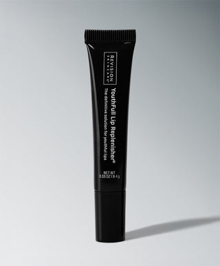 A black tube of Revision Skincare's YouthFull Lip Replenisher® stands upright on a light gray surface with soft lighting, displaying the product name and details in white text.