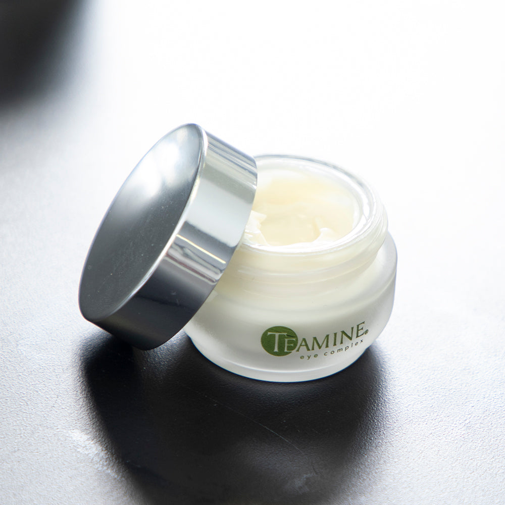 A small glass jar of Revision Skincare Teamine® Eye Complex 0.5 oz, with a silver lid to the side, reveals the light cream inside. Designed to target dark circles and fine lines, the jar sits on a smooth, dark surface.